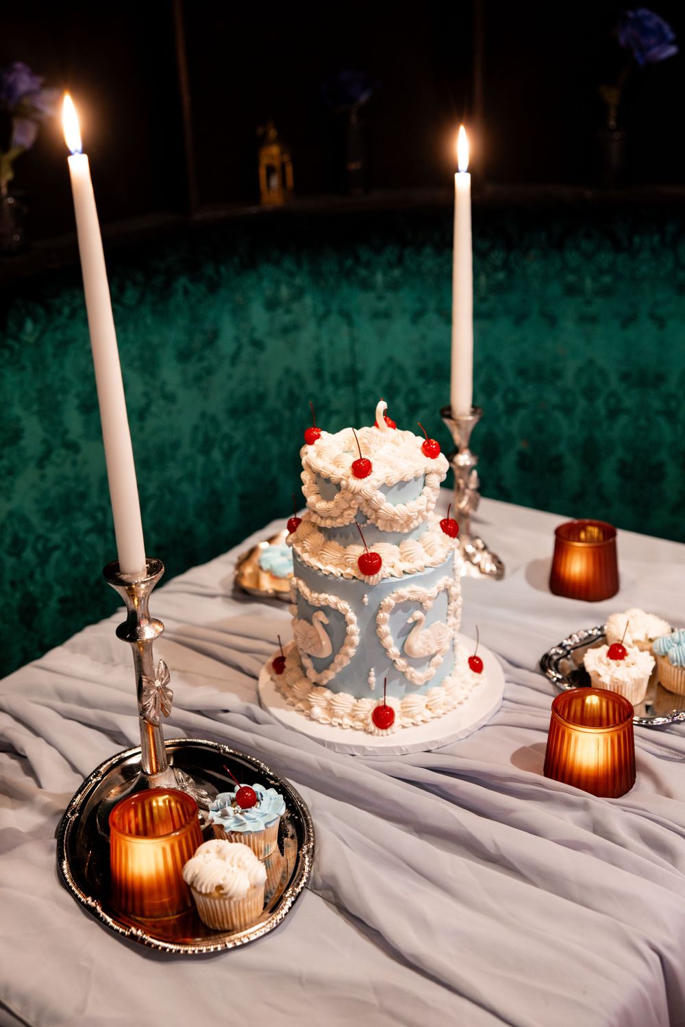 Two-tier blue wedding cake decorated with red cherries and white frosting, flanked by tall white candles on a draped table.
