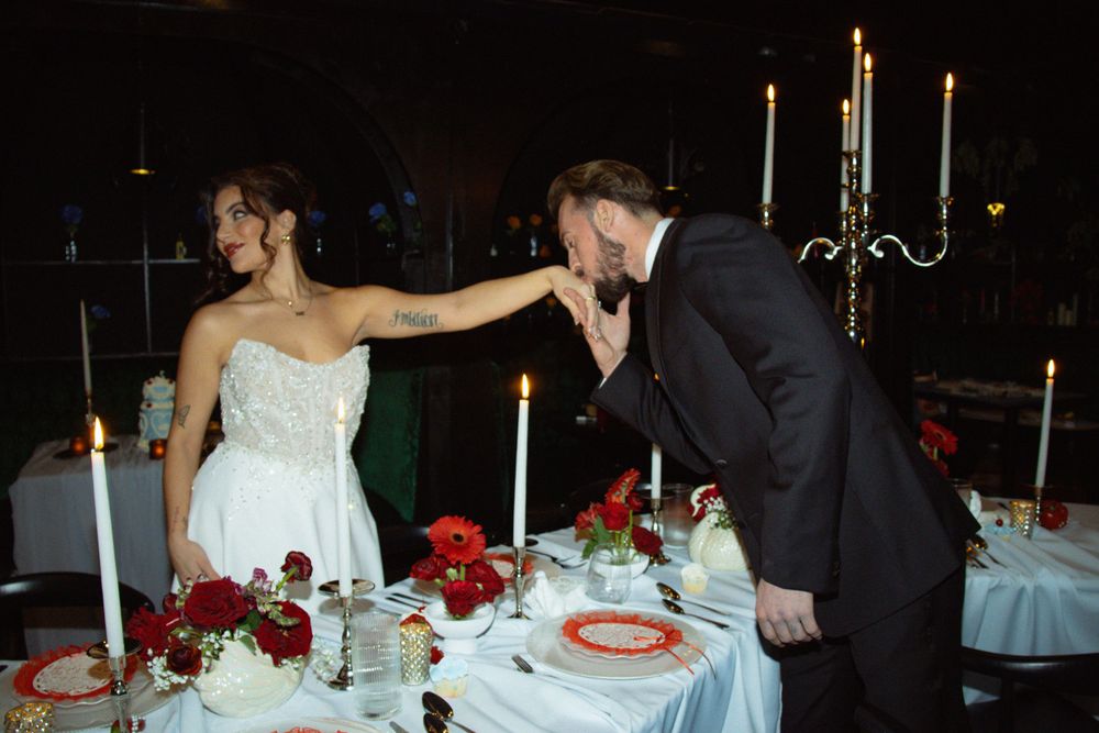 Bride in white strapless gown extending hand to groom in black suit at candlelit wedding reception table with red roses.