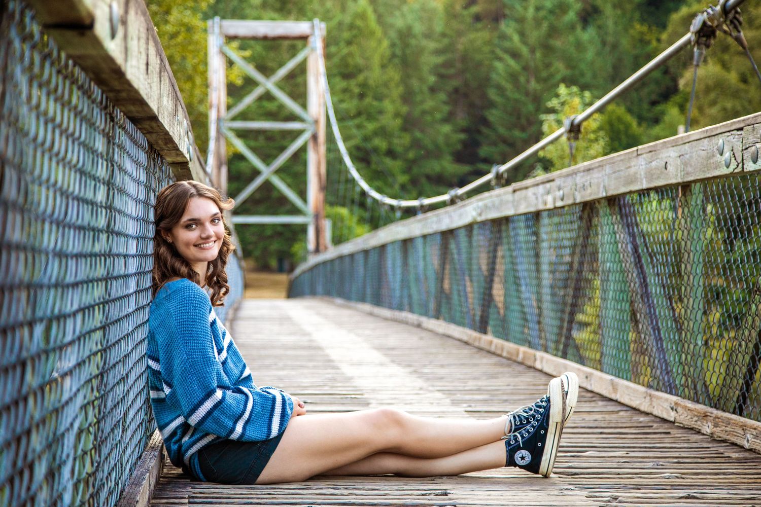 Person sitting on wooden suspension bridge surrounded by lush forest scenery.