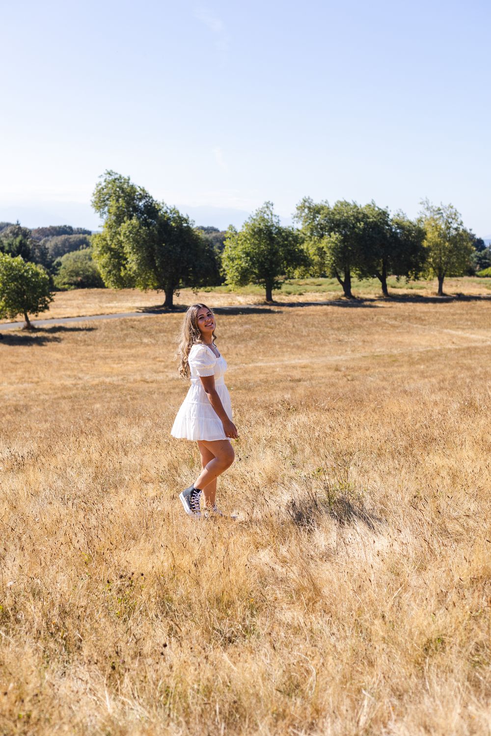 A person in a white dress walks through a golden summer field with scattered oak trees on a hillside.
