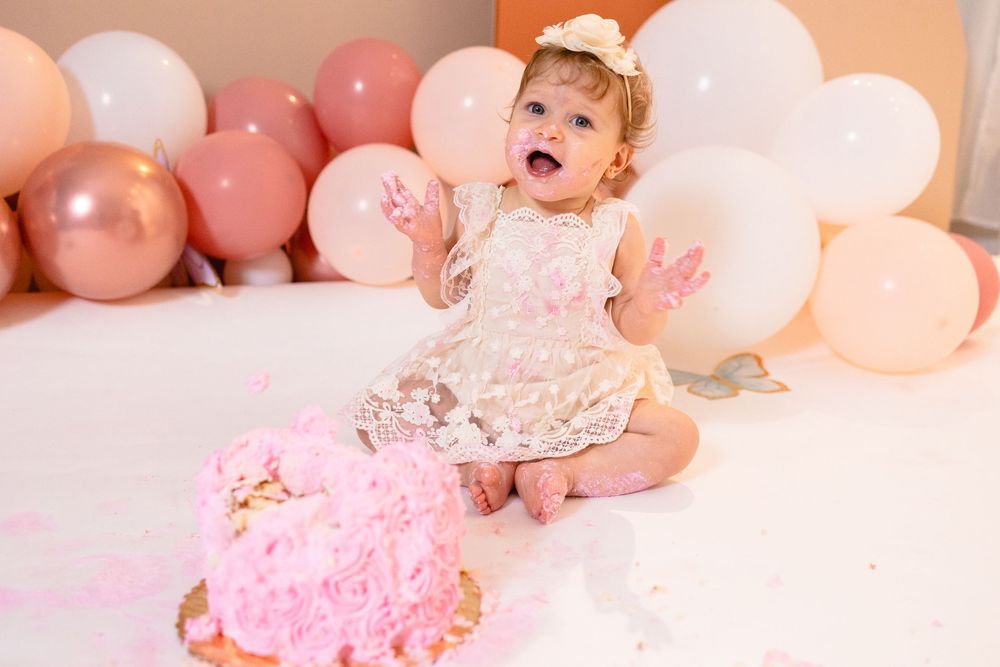 A baby in a lace outfit poses with pink balloons and cake during a first birthday cake smash photo session.