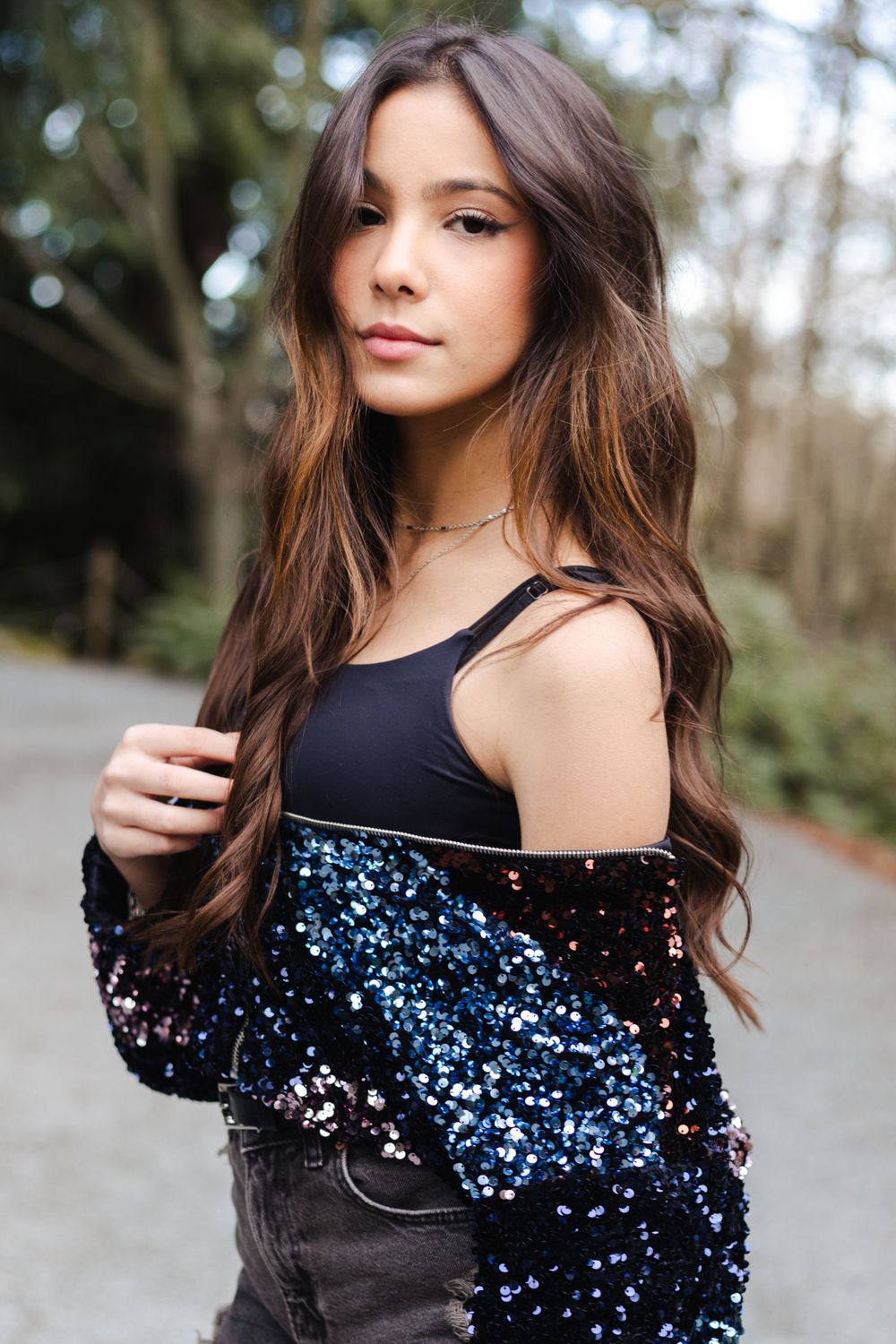 A model wearing a black top with sparkly blue sequin details poses against a natural outdoor background.