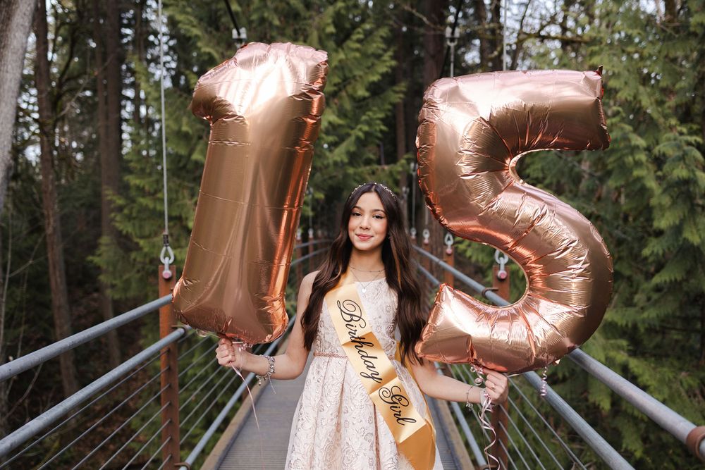 A person in a white dress holds rose gold number balloons in a forest setting during golden hour.