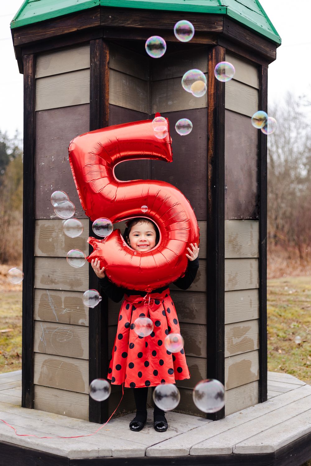 A child in a red number 5 costume and polka dot dress stands by a wooden structure while bubbles float in the air.