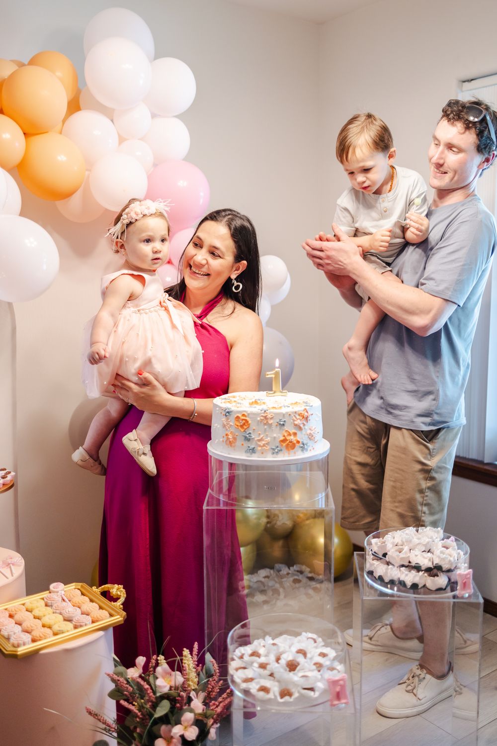 A family celebrates at a baby's first birthday party with pink and orange balloons and dessert table decorations.
