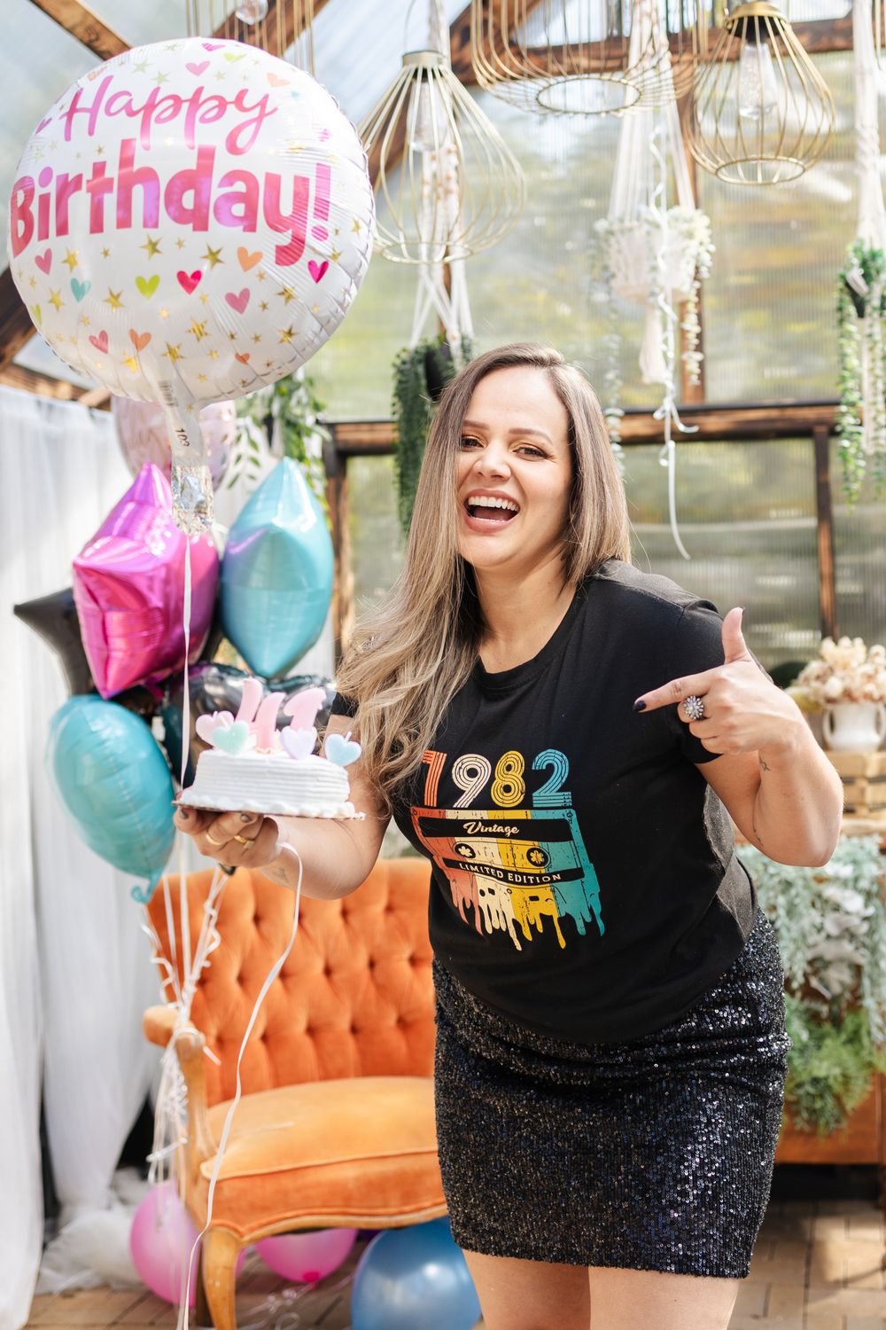 Birthday celebration with sparkly dress, vintage orange couch and colorful party balloons in a bright indoor setting.