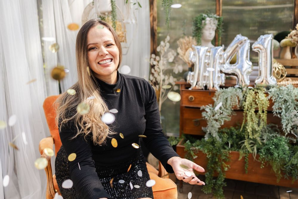 A person in black clothing smiles while working in a plant nursery with potted flowers and greenery.