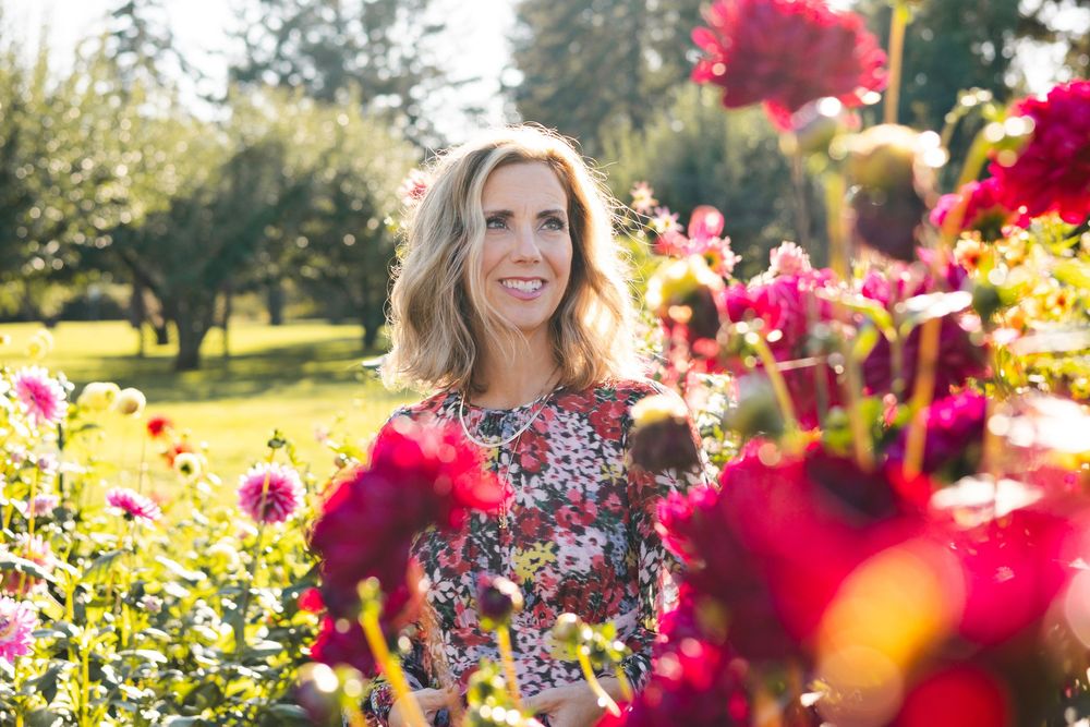 A person stands among vibrant red dahlias in a sunny garden with trees in the background.