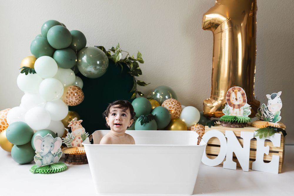 Safari themed first birthday party decorations with balloon garland, stuffed animals and gold lettering on white wall.