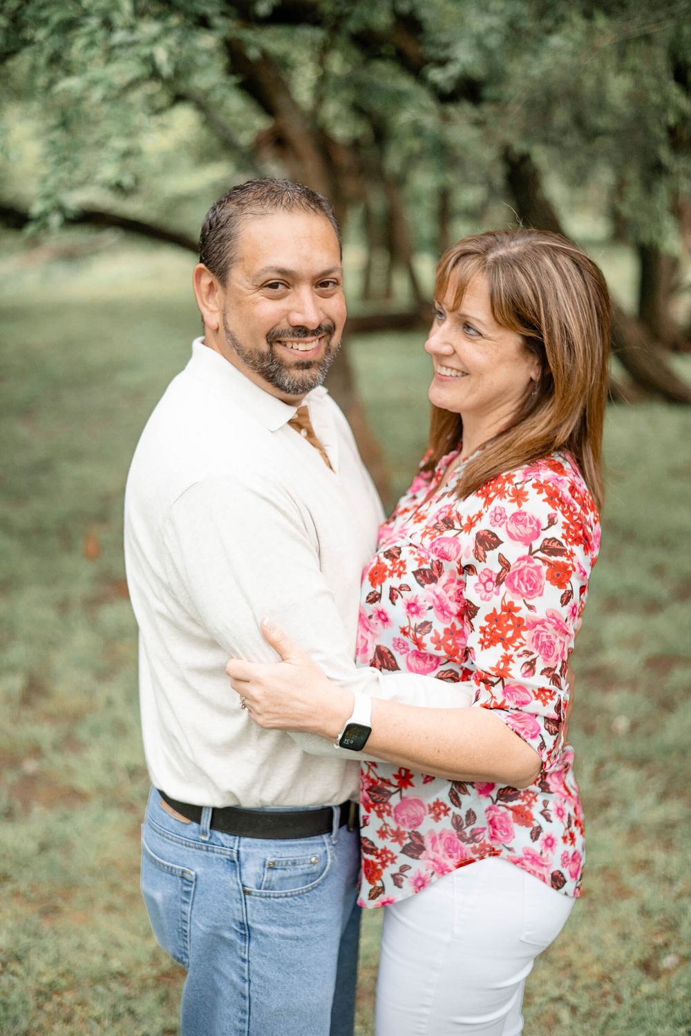 A couple embraces outdoors in a park setting, with her wearing a floral pink top and him in a white shirt and jeans.