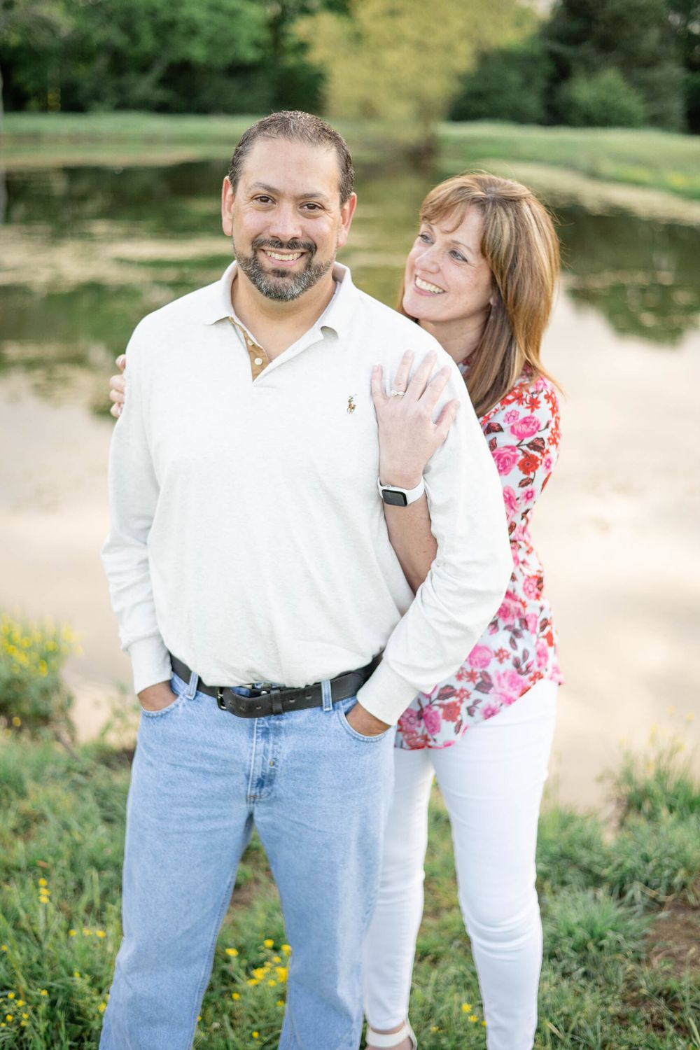 A couple embraces outdoors near a pond on a sunny day, with the woman wearing a floral top and white pants.