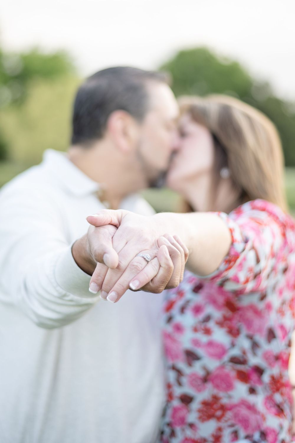 A romantic engagement photo showing clasped hands with a ring while a couple shares a tender kiss in soft focus.