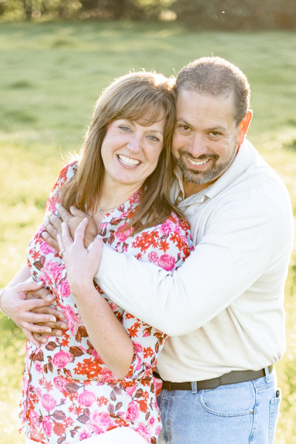 A couple embraces and smiles together outdoors in a sunlit field, with her wearing a pink floral dress and him in white.