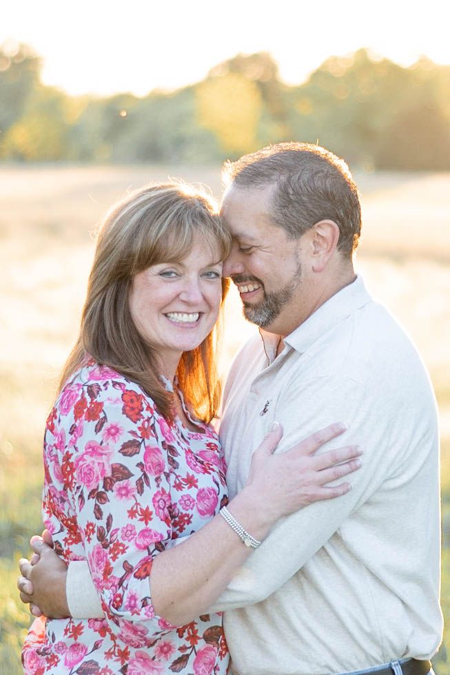 Happy couple embraces in a sunlit field during sunset wearing floral pink top and cream sweater.