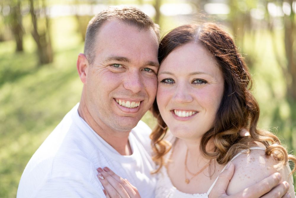 A couple poses together outdoors on a sunny day with a natural blurred forest background.