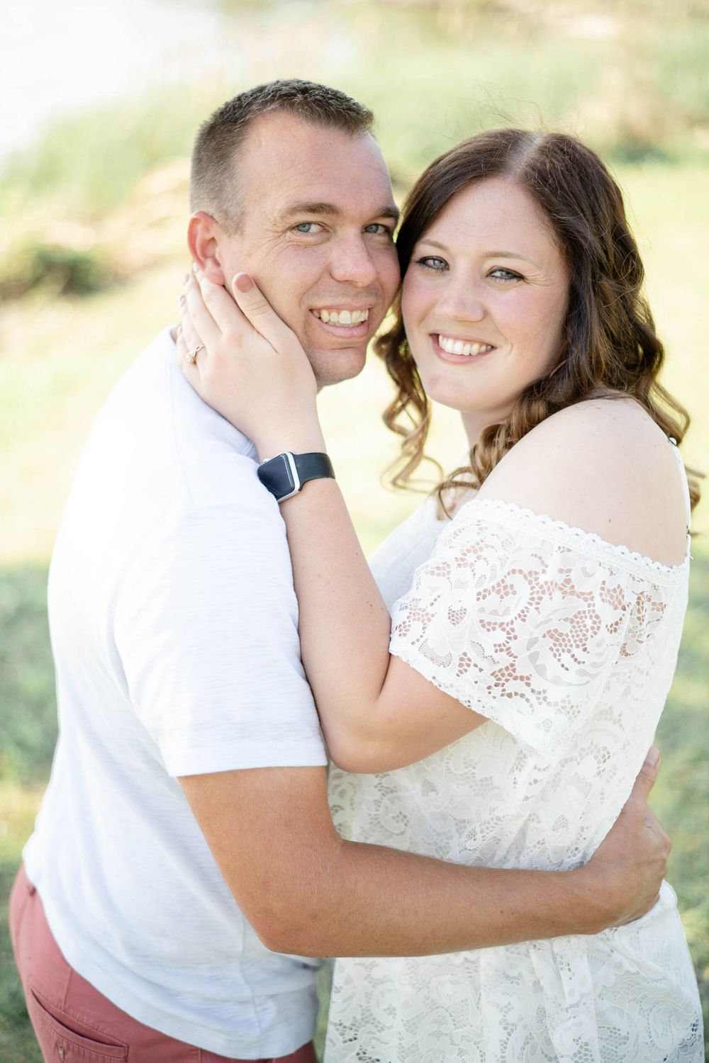 A romantic outdoor portrait of a couple embracing, with the woman wearing a white lace dress in soft natural lighting.