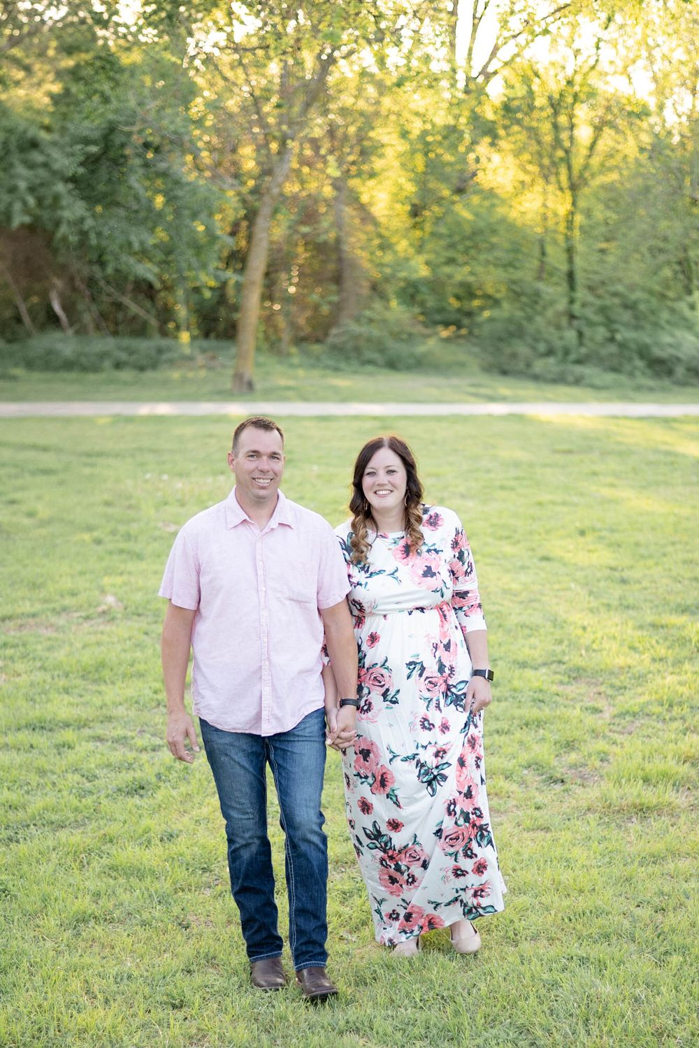 A couple walks together on green grass in a park during golden hour with trees in the background.