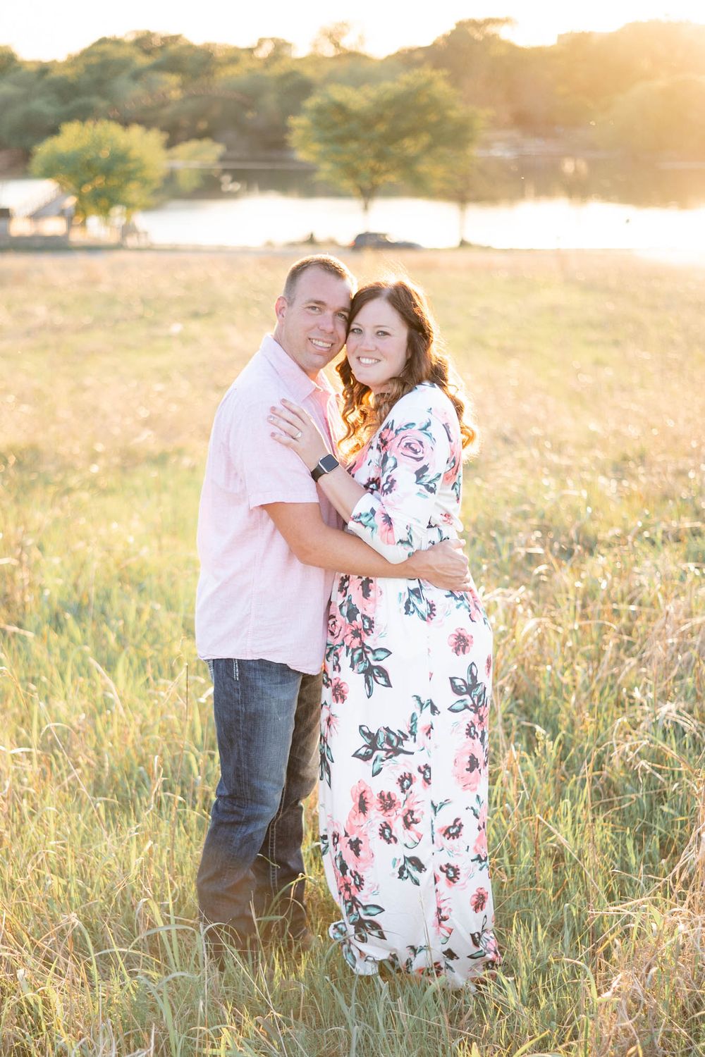 A couple embraces in a sunny meadow at golden hour, with her wearing a floral maxi dress against a natural landscape.