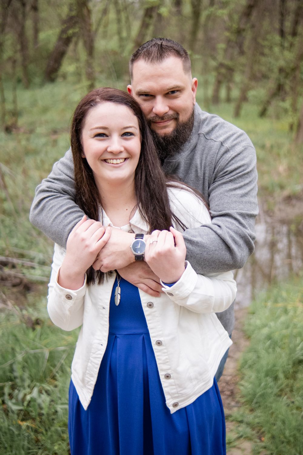 A couple embraces outdoors in a wooded area, with her wearing a blue dress and white jacket against a natural backdrop.