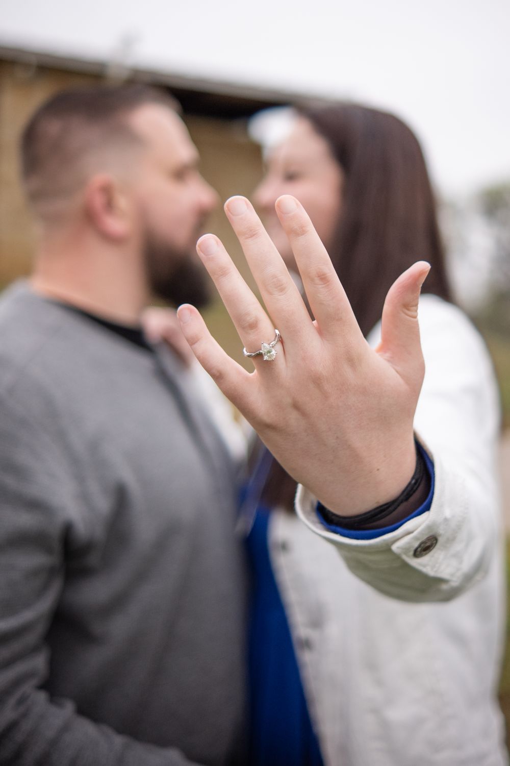 A close-up shot of a hand displaying an engagement ring with a couple embracing in soft focus in the background.