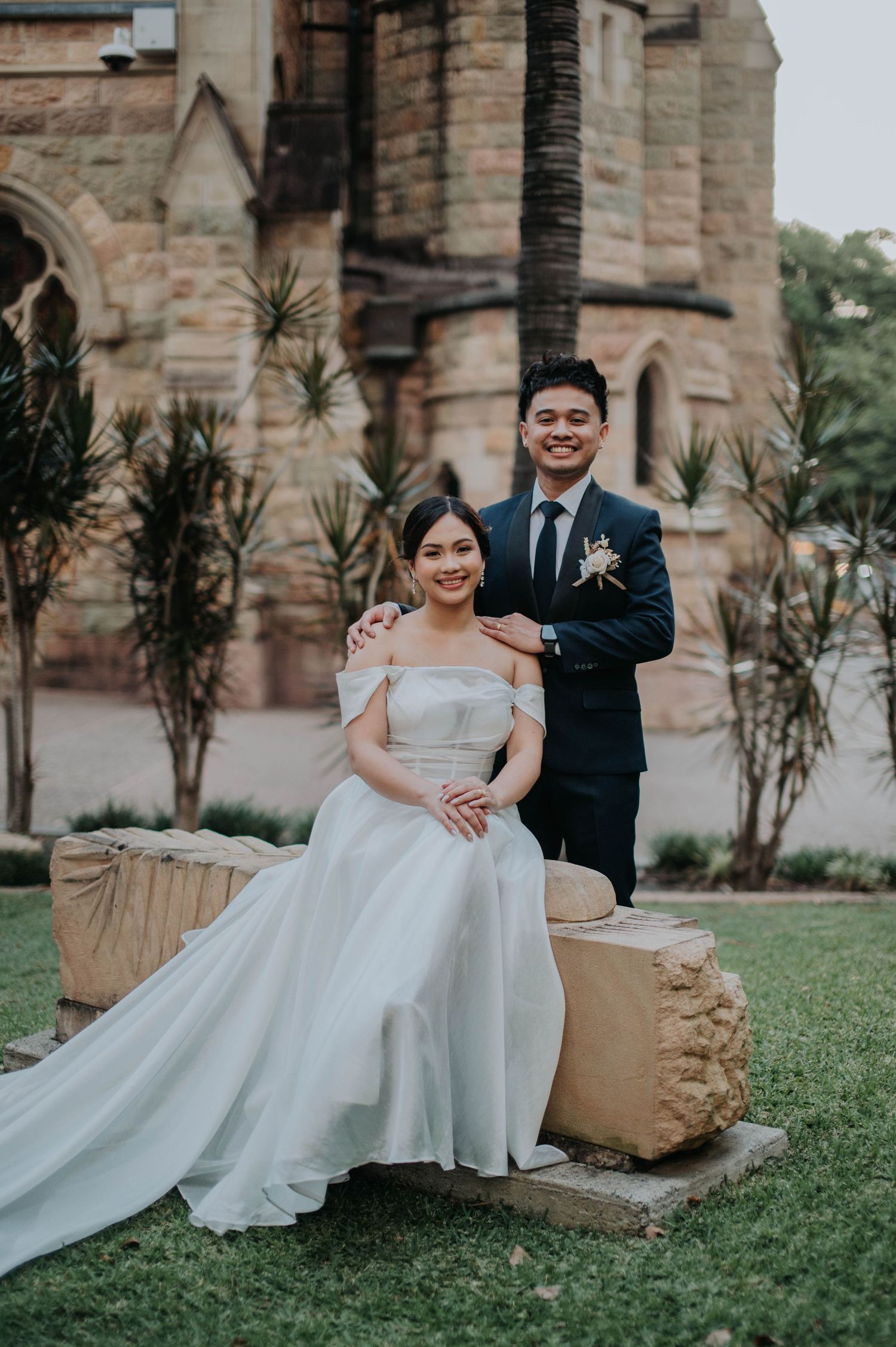 A couple poses for portraits on a garden bench outside a Gothic stone church.