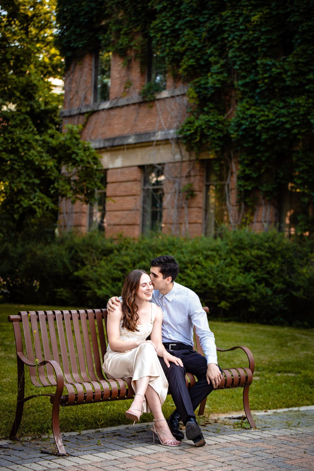 A couple shares a tender moment on a bench outside an ivy-covered brick building.
