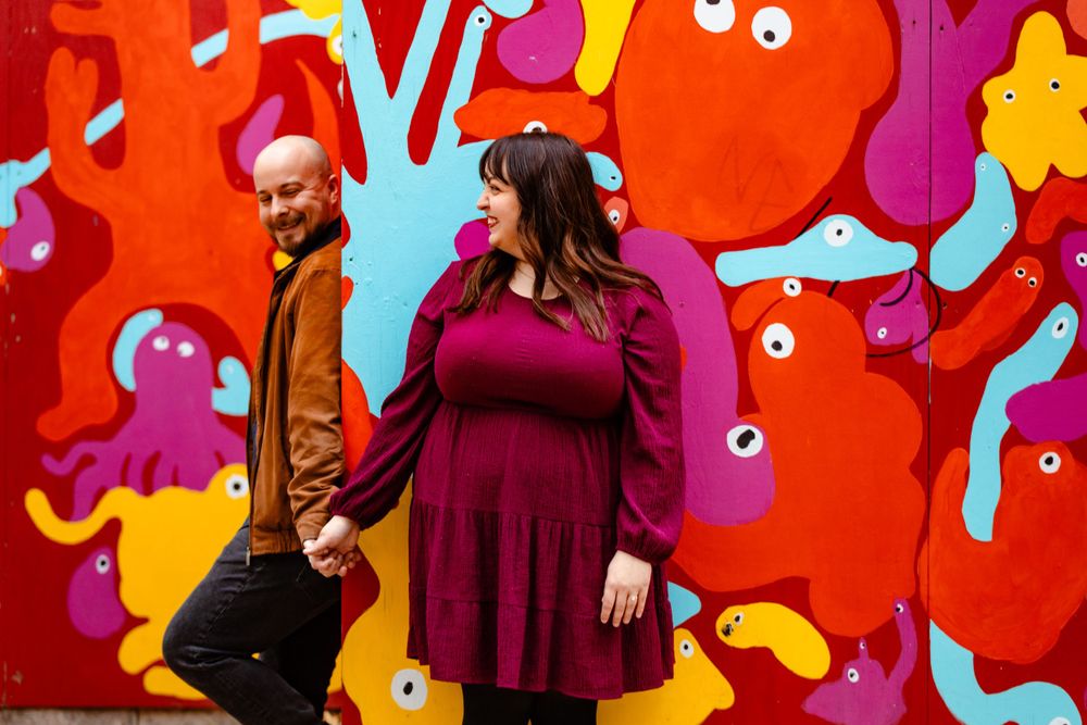 A couple poses playfully against a bright red wall decorated with whimsical cartoon characters.