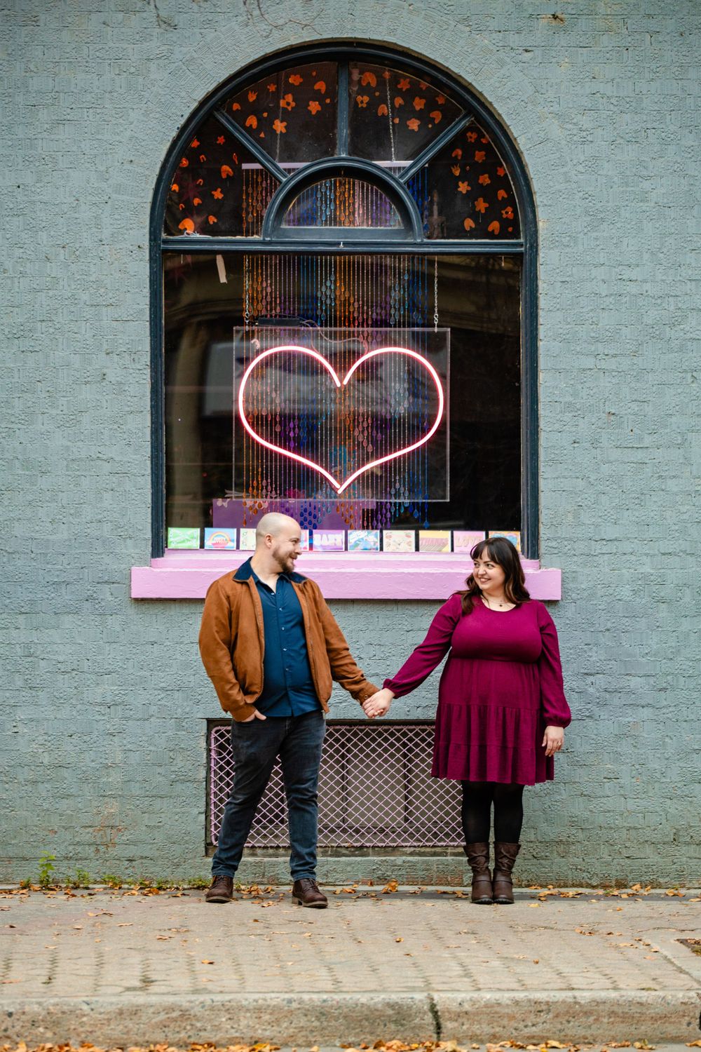 A couple stands in front of a wall with a neon heart sign and arched window decor.