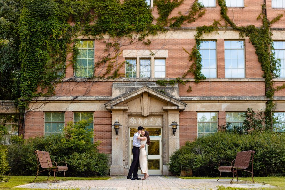 A couple shares an intimate moment in front of a historic brick building with ivy.