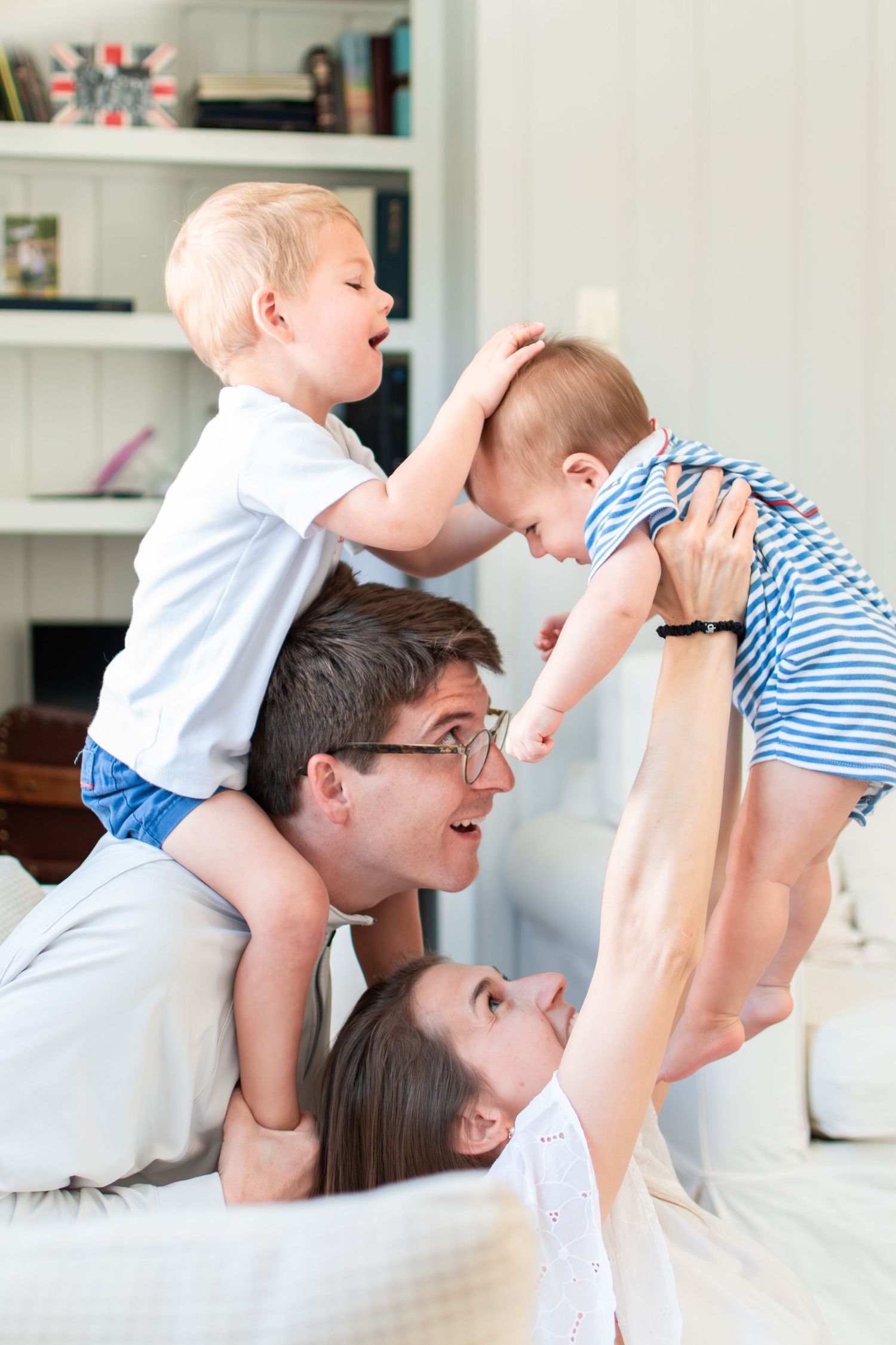 Two adults lie on a white couch playing and lifting two toddlers up in a playful moment at home.