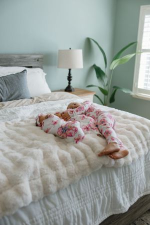 Sleeping infant wearing pink floral pajamas lying on white bedding in a mint green bedroom with a tall plant.