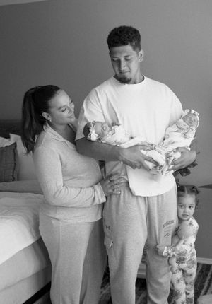 Black and white family photo showing parents holding a newborn baby with a toddler standing nearby in a bedroom setting.