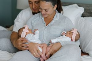 Two people in gray lounge clothes hold and gaze lovingly at twin infants on a bed.