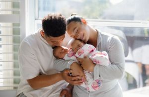 A couple in white clothing cuddles with their newborn baby near a window, filled with soft natural light.