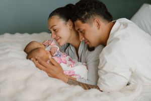 Two parents lovingly embrace their sleeping newborn baby while resting together on white bedding.
