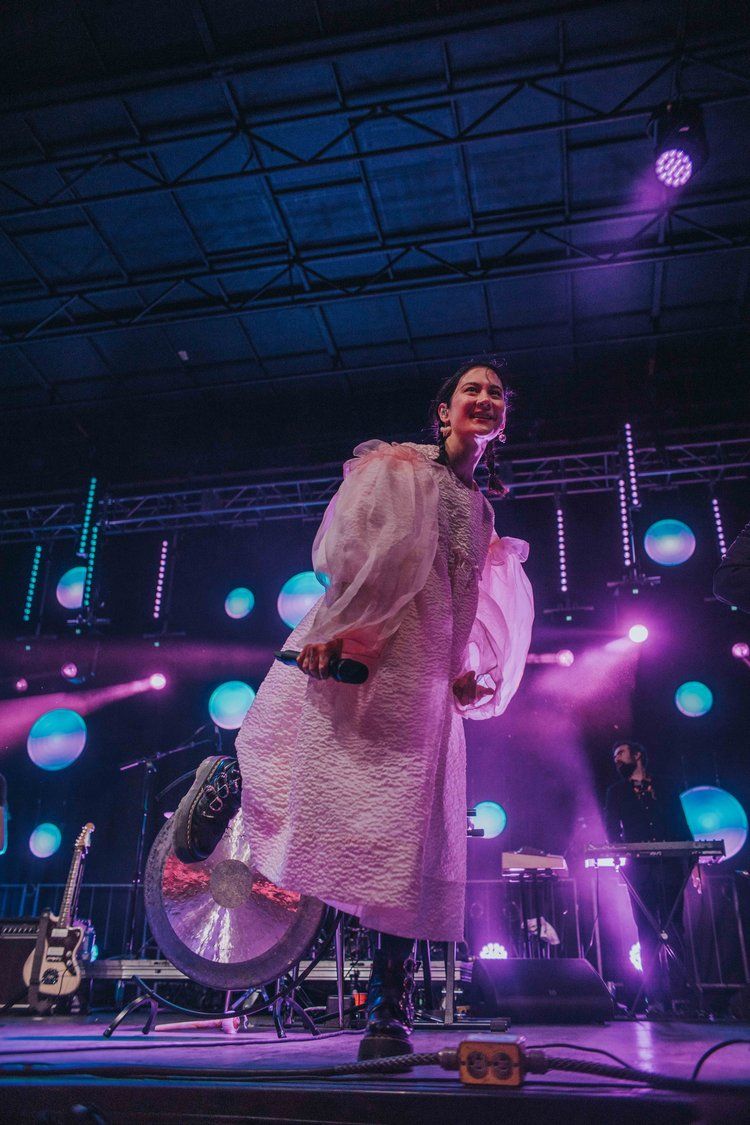 Concert performer in white textured coat stands on stage with dramatic purple and blue lighting and illuminated orbs in background.