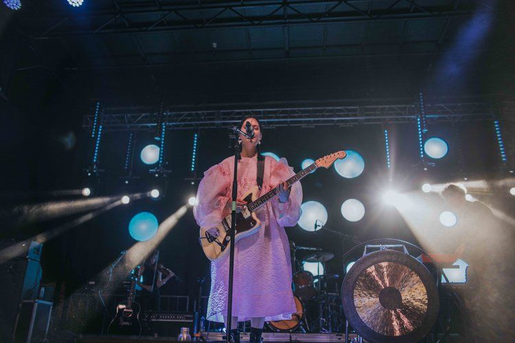 Concert performer in a pink dress plays guitar on stage with bright spotlights and lighting effects in the background.
