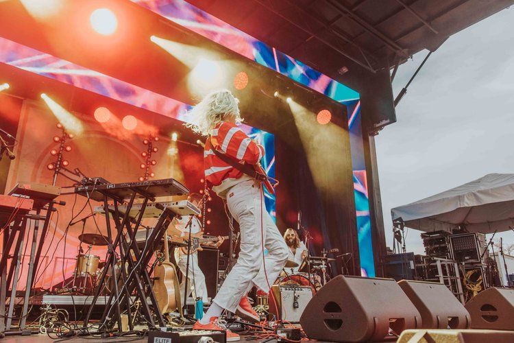 A performer in a white outfit jumps energetically on stage during a concert with colorful lighting and musical equipment.