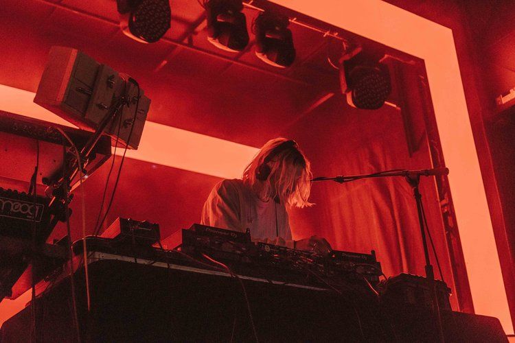 DJ performs behind decks at a concert with dramatic red stage lighting casting shadows.