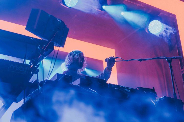 An electronic musician performs on stage with synthesizers against vibrant blue and orange lighting effects.