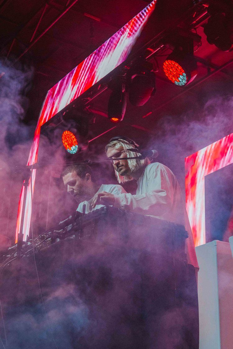DJs perform at an electronic music concert with dramatic red lighting and atmospheric smoke effects on stage.
