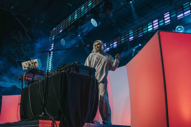 Electronic music performer on stage with red illuminated backdrop and digital equipment at a concert venue.