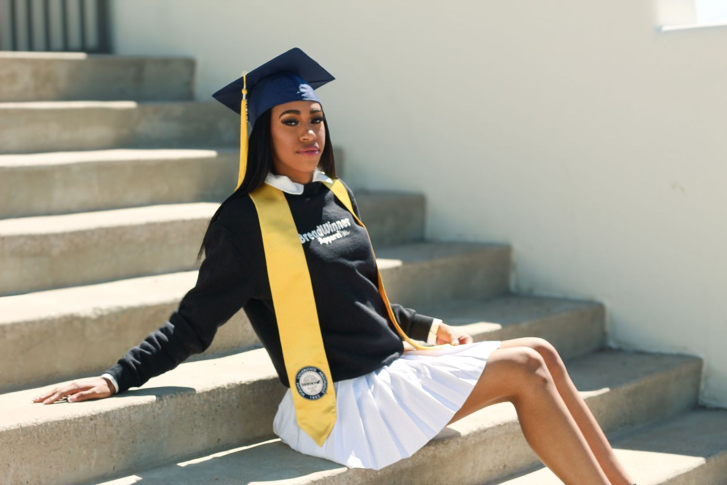 Graduate in cap and gown sitting on concrete steps in bright sunlight.