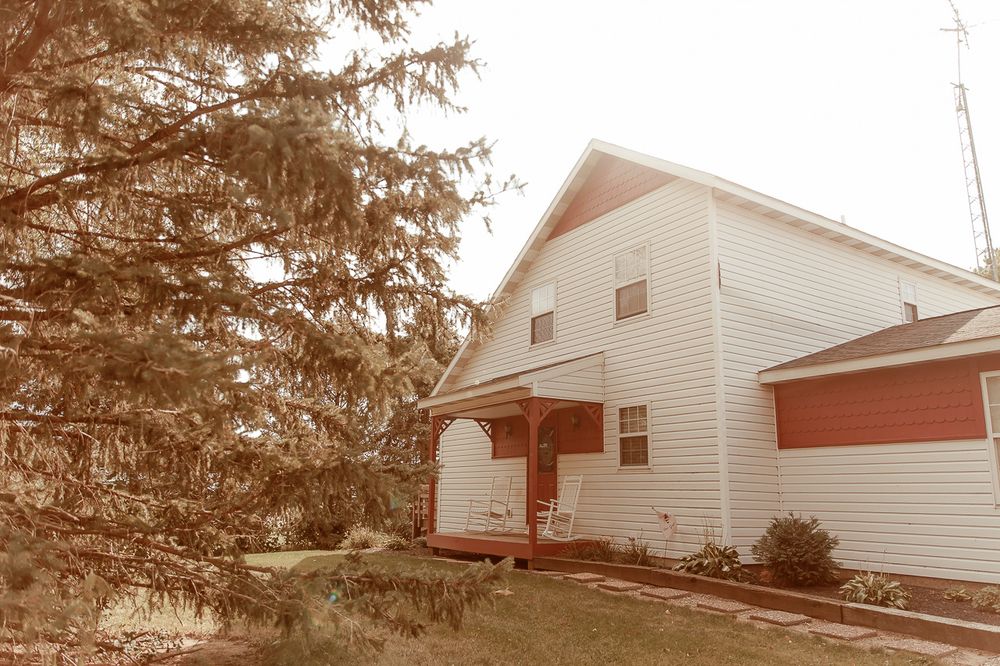 Vintage sepia-toned photo of a traditional white two-story farmhouse with covered porch and evergreen tree in foreground.