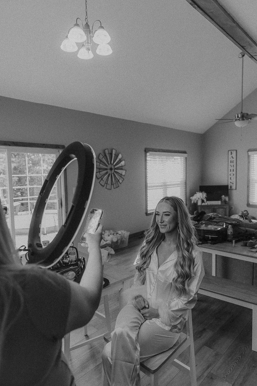 Behind the scenes black and white photo of bridal preparation with ring light and photographer in home setting.