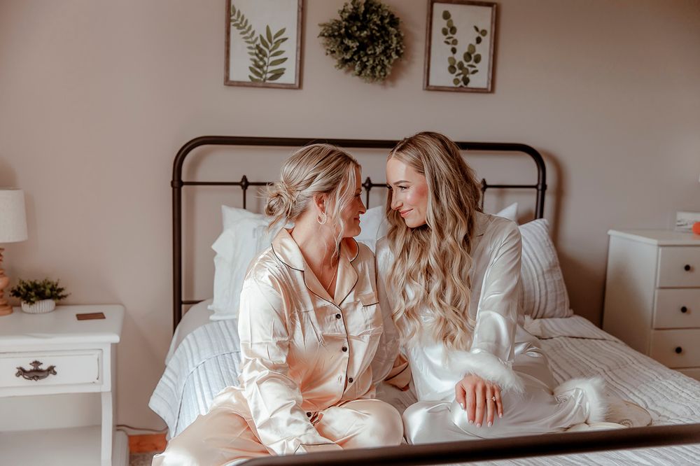 Two friends in white sleepwear share a cozy moment while sitting on a bed with rustic decor in the background.