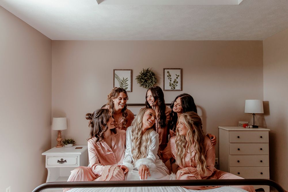 Five friends in matching sleepwear sit together on a bed, sharing laughter in a warmly decorated bedroom.