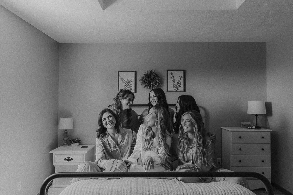A black and white photo showing a group laughing together on a bed with artwork and lamps in the background.