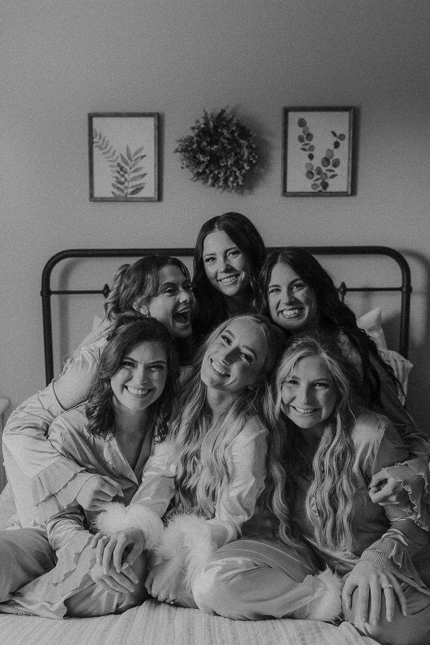 Black and white photo of friends in matching white outfits sitting together on a bed with botanical artwork on wall.