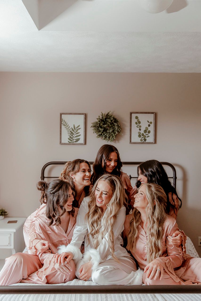 Group wearing matching pink satin robes and white pajamas sitting on a bed with botanical artwork in background.